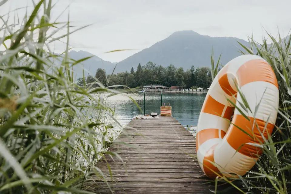 A wooden boardwalk on Lake Tegernsee with a lifebuoy, reeds, and a view of the surrounding mountains.