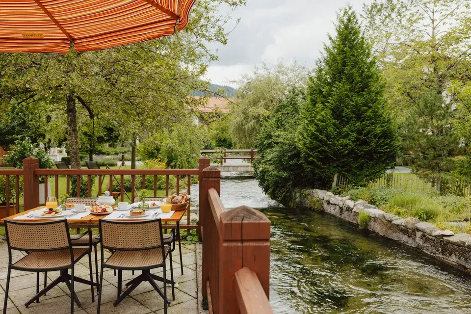 View from the terrace, where a breakfast table is set, overlooking a stream lined with trees and shrubs.