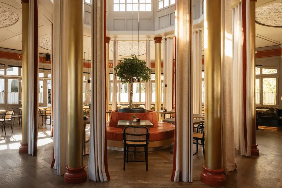 Dining area at Bachmair Weissach featuring tall columns, curtains, and seating alcoves—a light-filled pavilion with a tree in the center