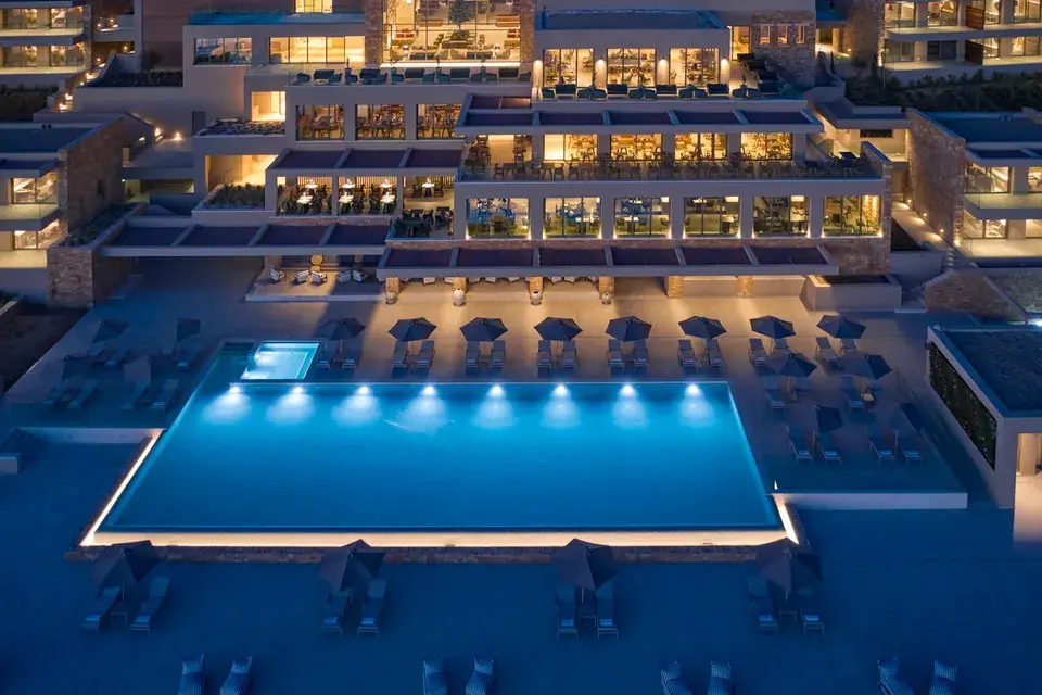 Elix - An illuminated outdoor pool with lounging areas and buildings in the background at night.