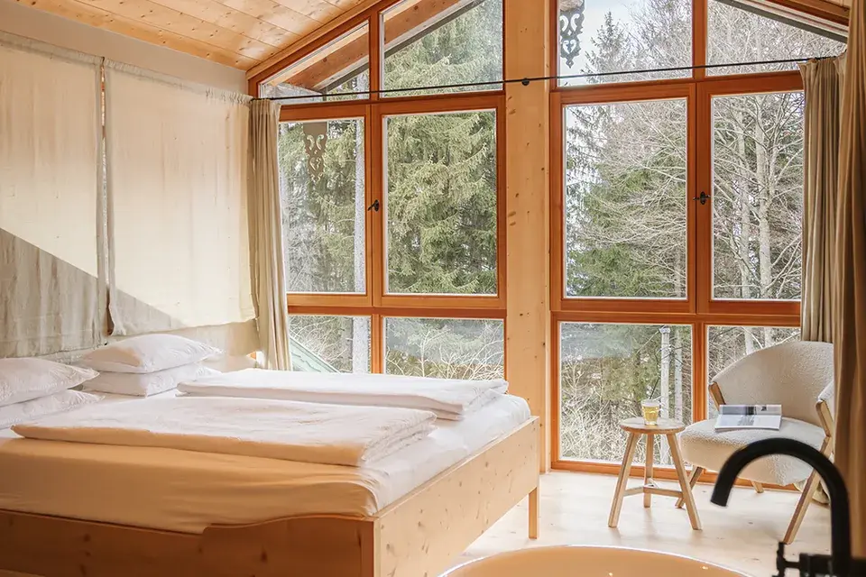 Bedroom of a chalet suite at the Chiemsee Chalet, featuring a freestanding bathtub, large wooden windows, and a view of the countryside.
