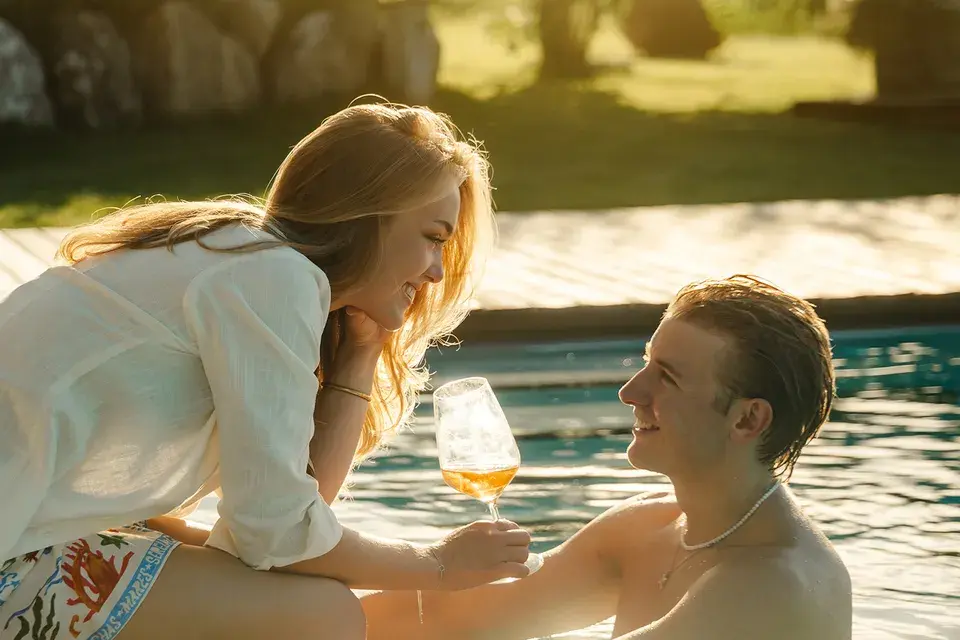 Frau sitzt am Pool mit Glas in der Hand und beugt sich herunter zu Mann im Pool im Sonnenschein.