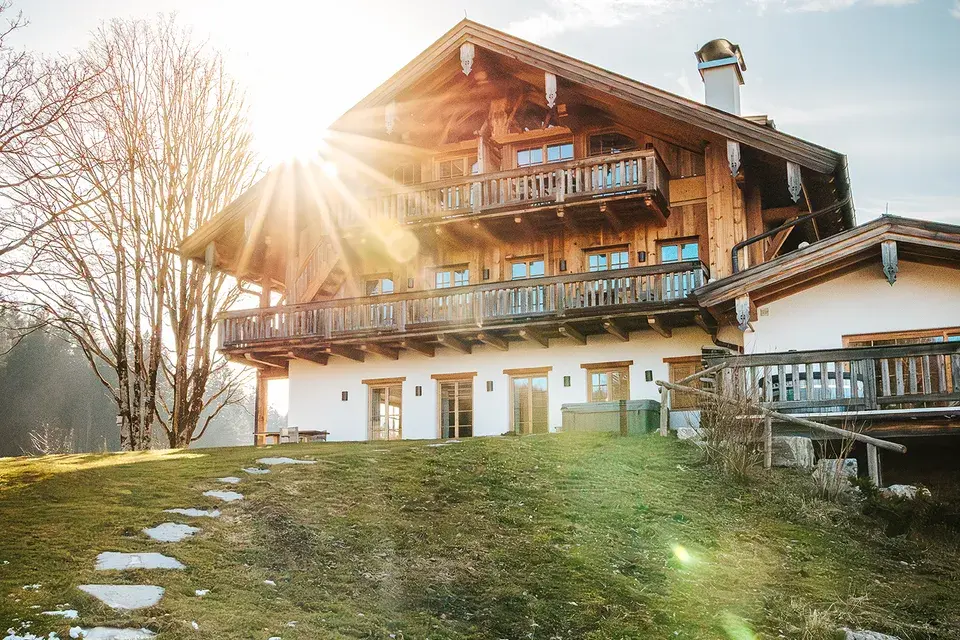 Exterior view of the Chiemsee Chalet with wooden balconies, a terrace, a garden, and rays of sunshine.