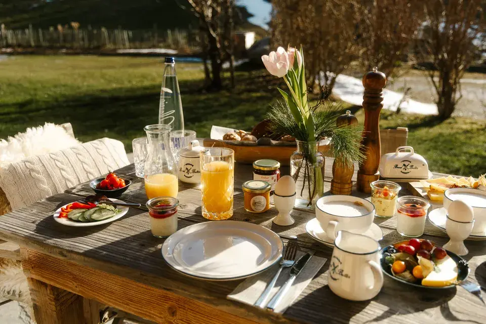 Outdoor breakfast table set with bread, fruit, eggs, juices, and other dishes.