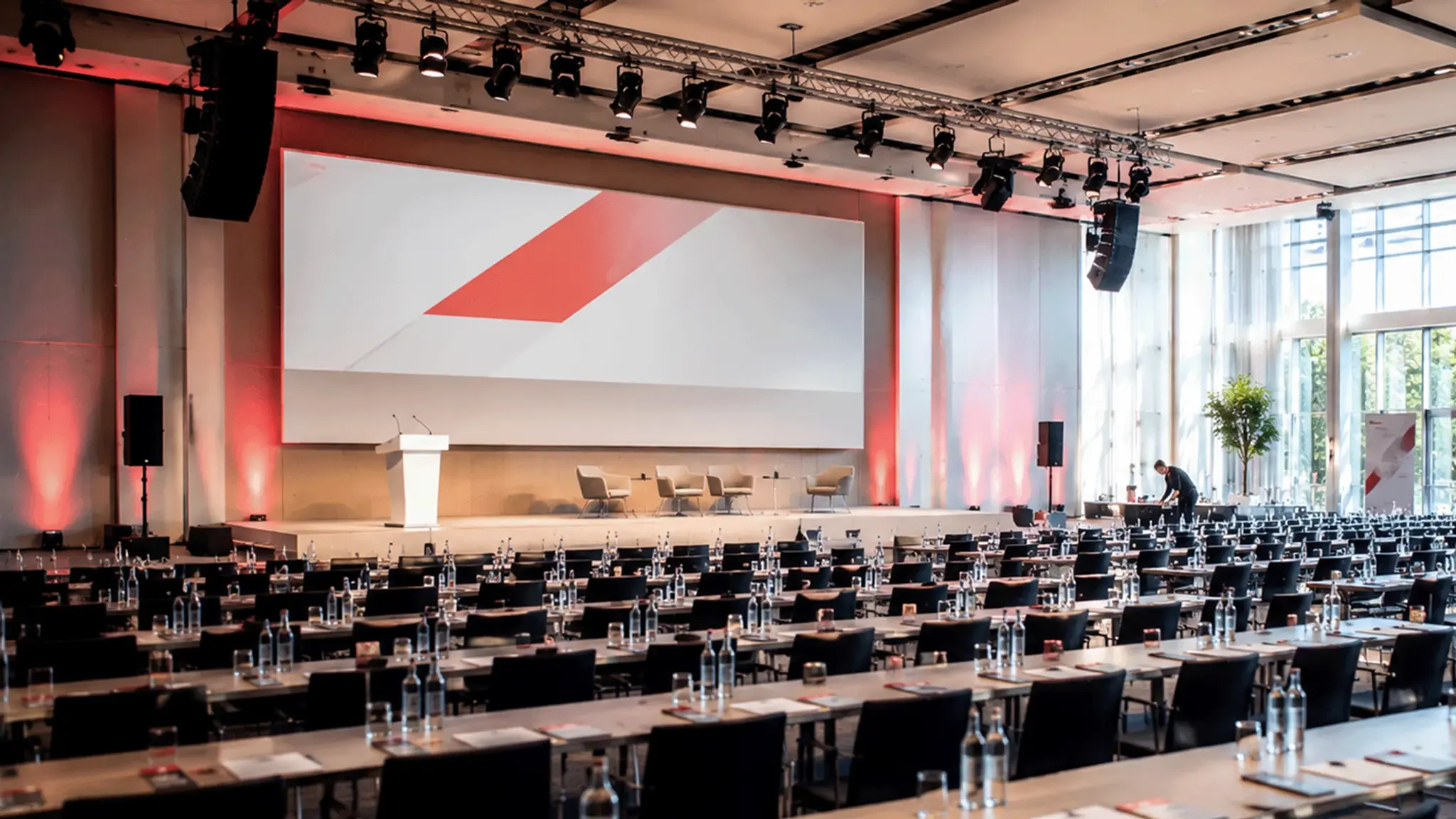 Großer Konferenzsaal mit Reihen aus Tischen und Stühlen, einer Bühne mit Rednerpult und einer großformatigen Präsentationsfläche.