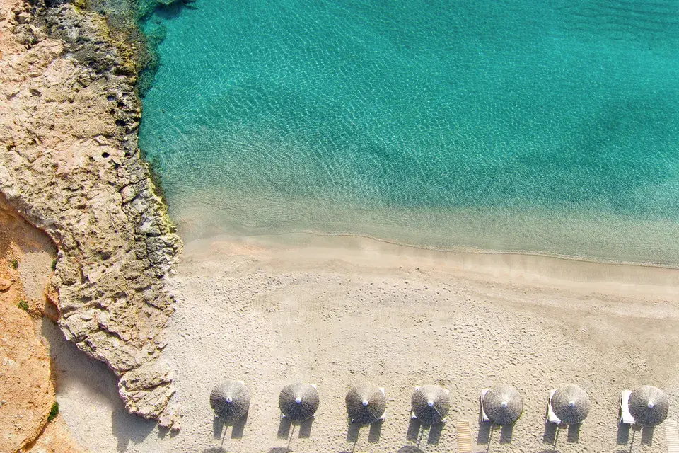 A bird's-eye view of a bright sandy beach, beach umbrellas, and lounge chairs, as well as the clear, turquoise sea in Daios Cove.