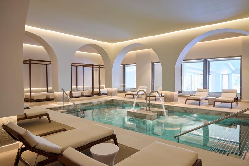 Indoor pool at the KĒPOS Spa at Daios Cove with lounge areas, large arches, and floor-to-ceiling windows overlooking the outdoors.