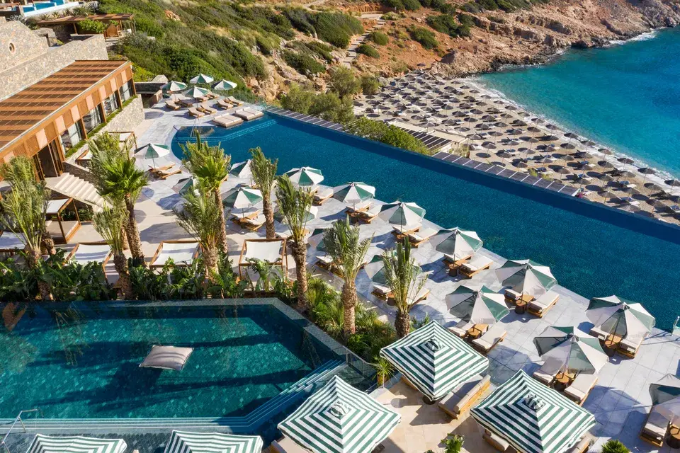 View across Daios Cove in Crete with a large infinity pool, sun terraces, and a private beach in a rocky bay by the sea.