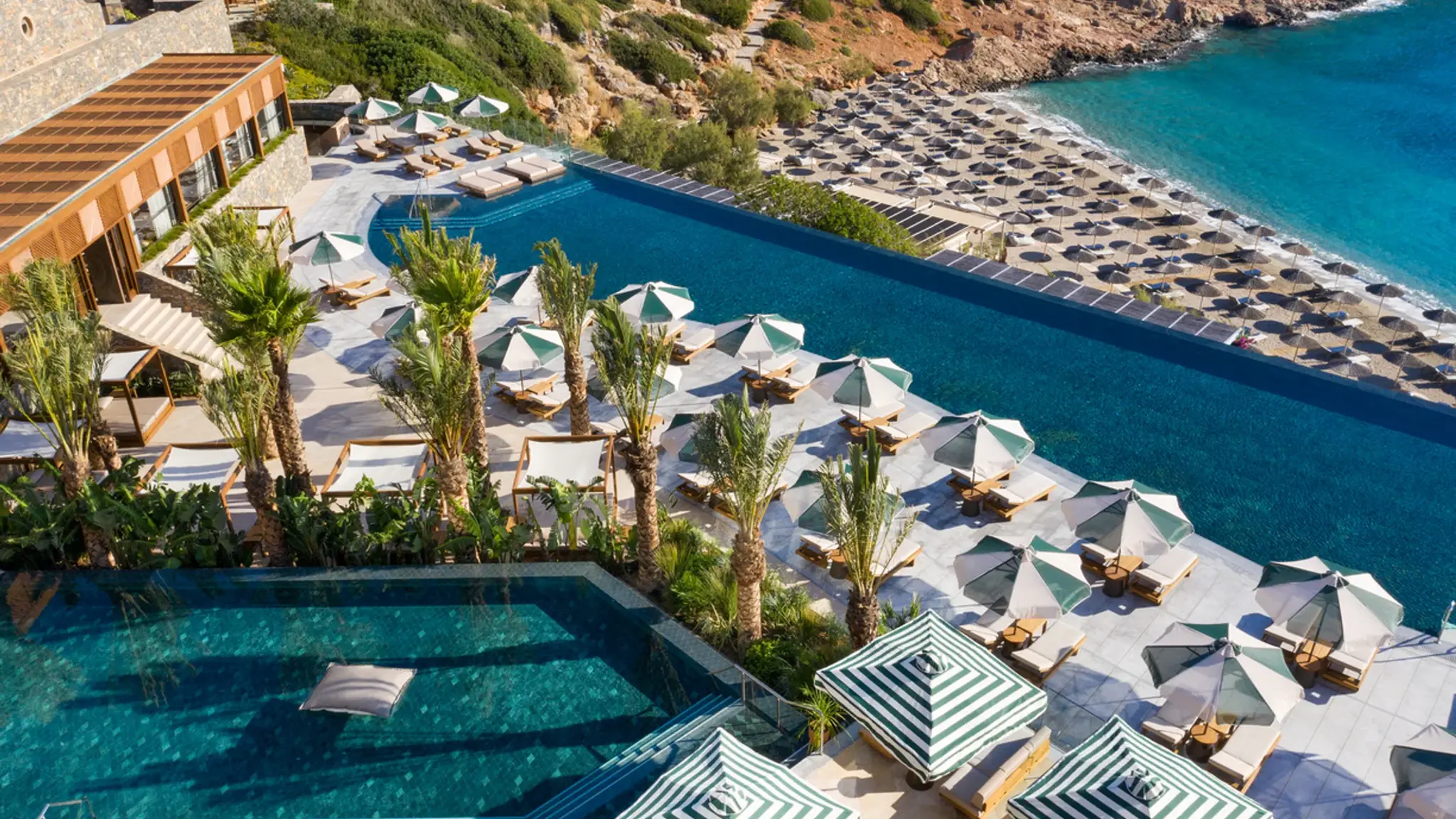 View across Daios Cove in Crete with a large infinity pool, sun terraces, and a private beach in a rocky bay by the sea.