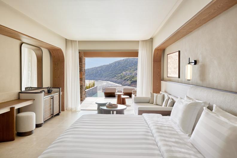 Bright bedroom at Daios Cove with a large bed, seating area, and open views of the terrace, sea, and opposite coastline.