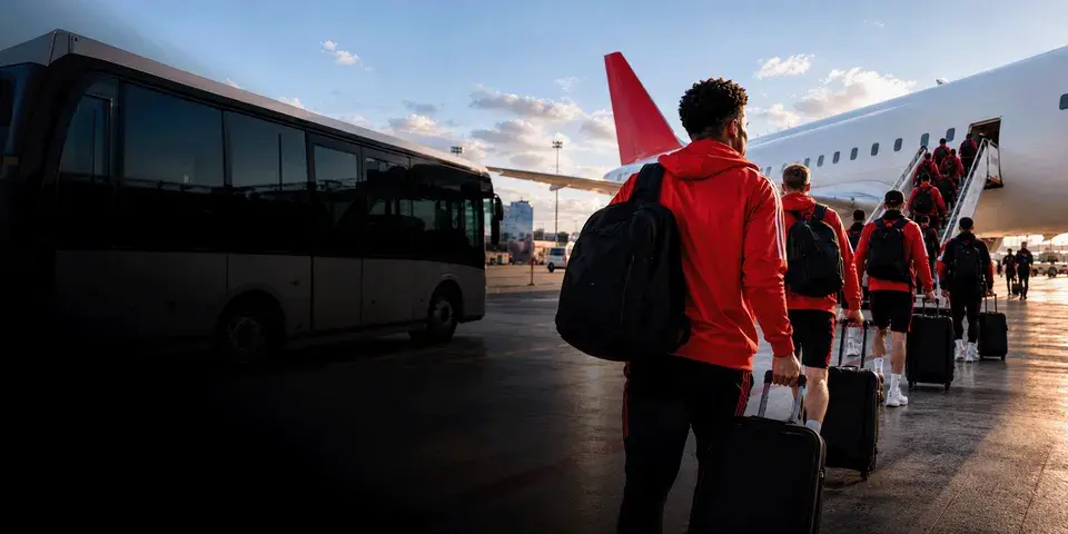 Mitglieder eines Sportteams gehen mit Rucksäcken und Rollkoffern über das Vorfeld zu einem Flugzeug und steigen über eine Treppe ein.