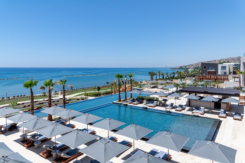 Infinity Pool über Eck mit Sonnenschirmen, Palmen und Liegen und Blick aufs Meer
