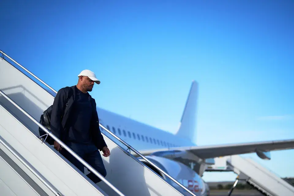 FC Bayern Trainer Vincent Kompany geht mit Rucksack und weißer Kappe die Treppe eines Flugzeugs hinunter.