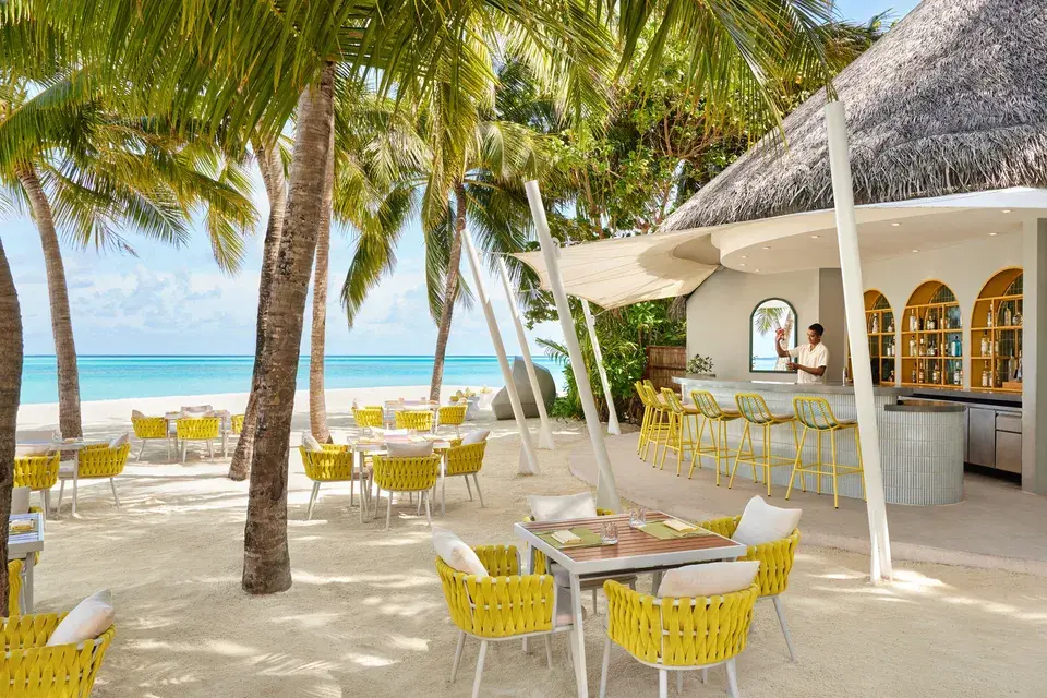 Open-air beachfront restaurant at Niyama Private Islands Maldives with tables and yellow chairs beneath palm trees overlooking the turquoise ocean.