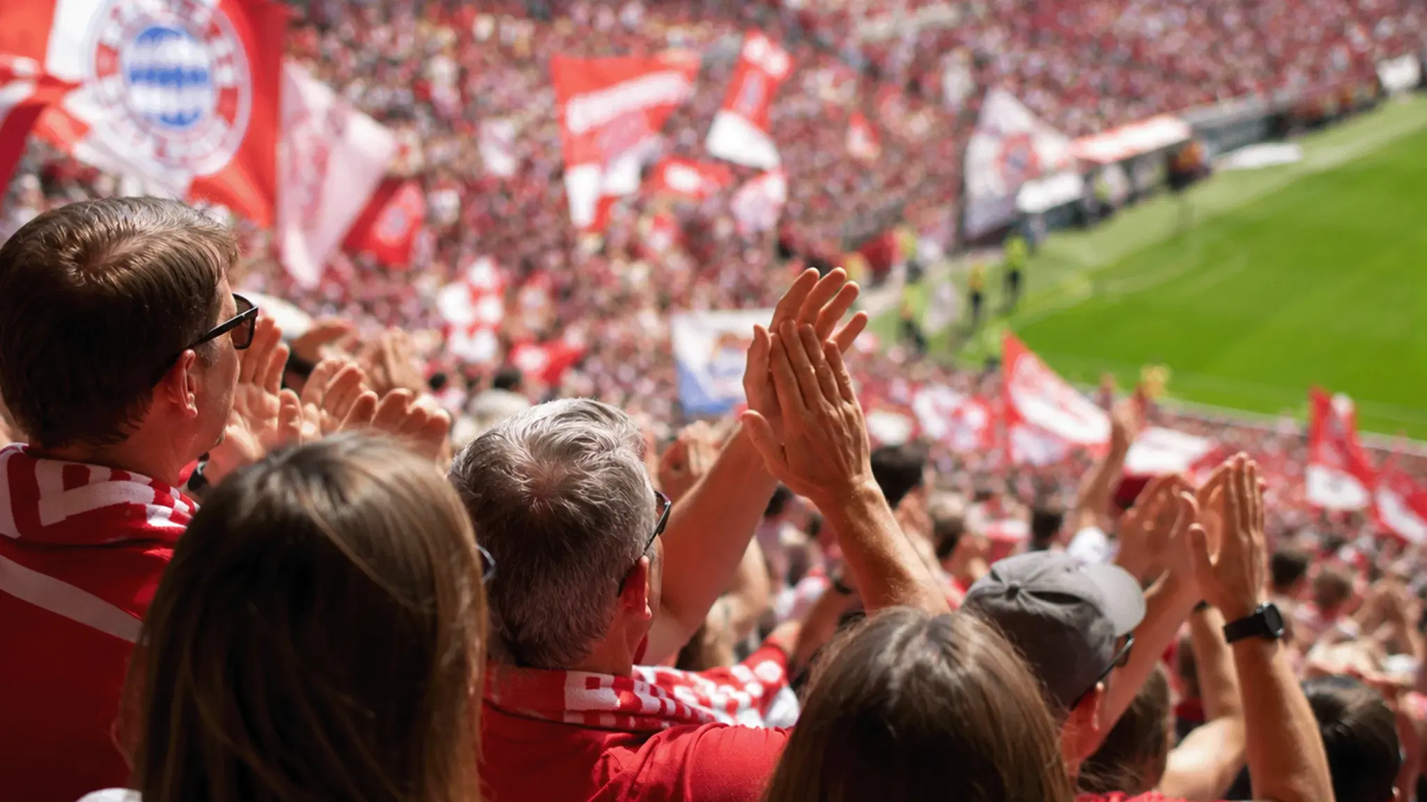 FC Bayern Fans klatschen Beifall auf einer voll besetzten Stadiontribüne, auf der zahlreiche rot-weiße Fahnen geschwenkt werden.