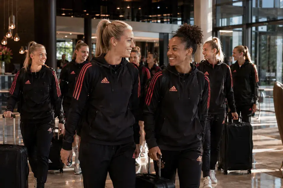 Spielerinnen eines Sportteams gehen in einheitlicher Sportkleidung mit Rollkoffern durch eine Hotellobby.