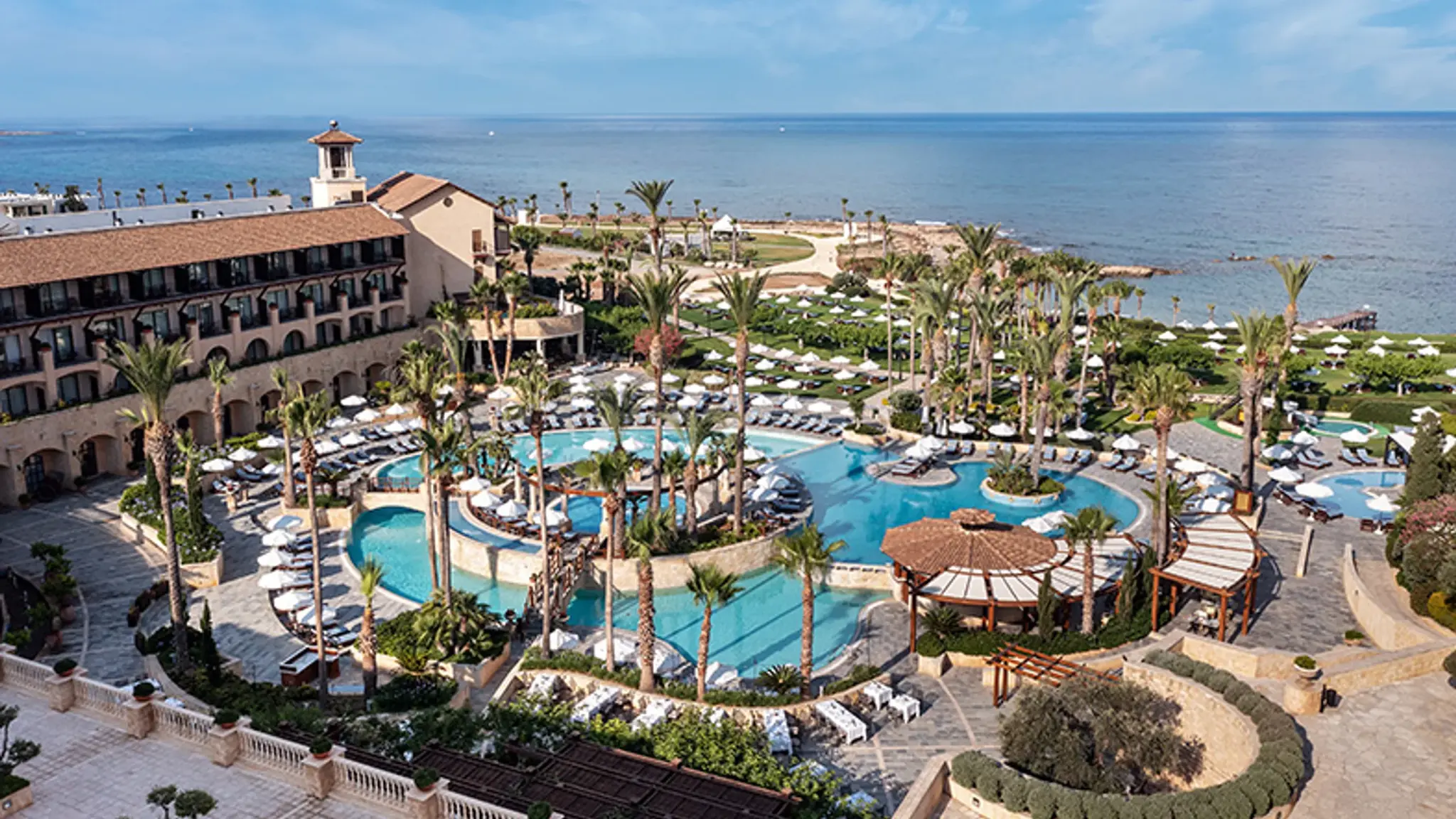 Elysium in Paphos with an expansive pool area, palm trees, and views of the Mediterranean Sea